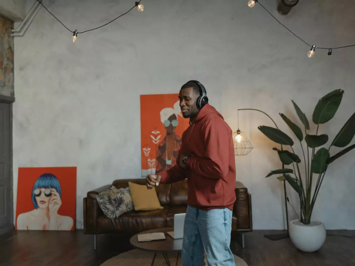 A man in a red jacket listens to music with headphones on, he is holding a book in his hands and he is smiling, he is wearing a cap.