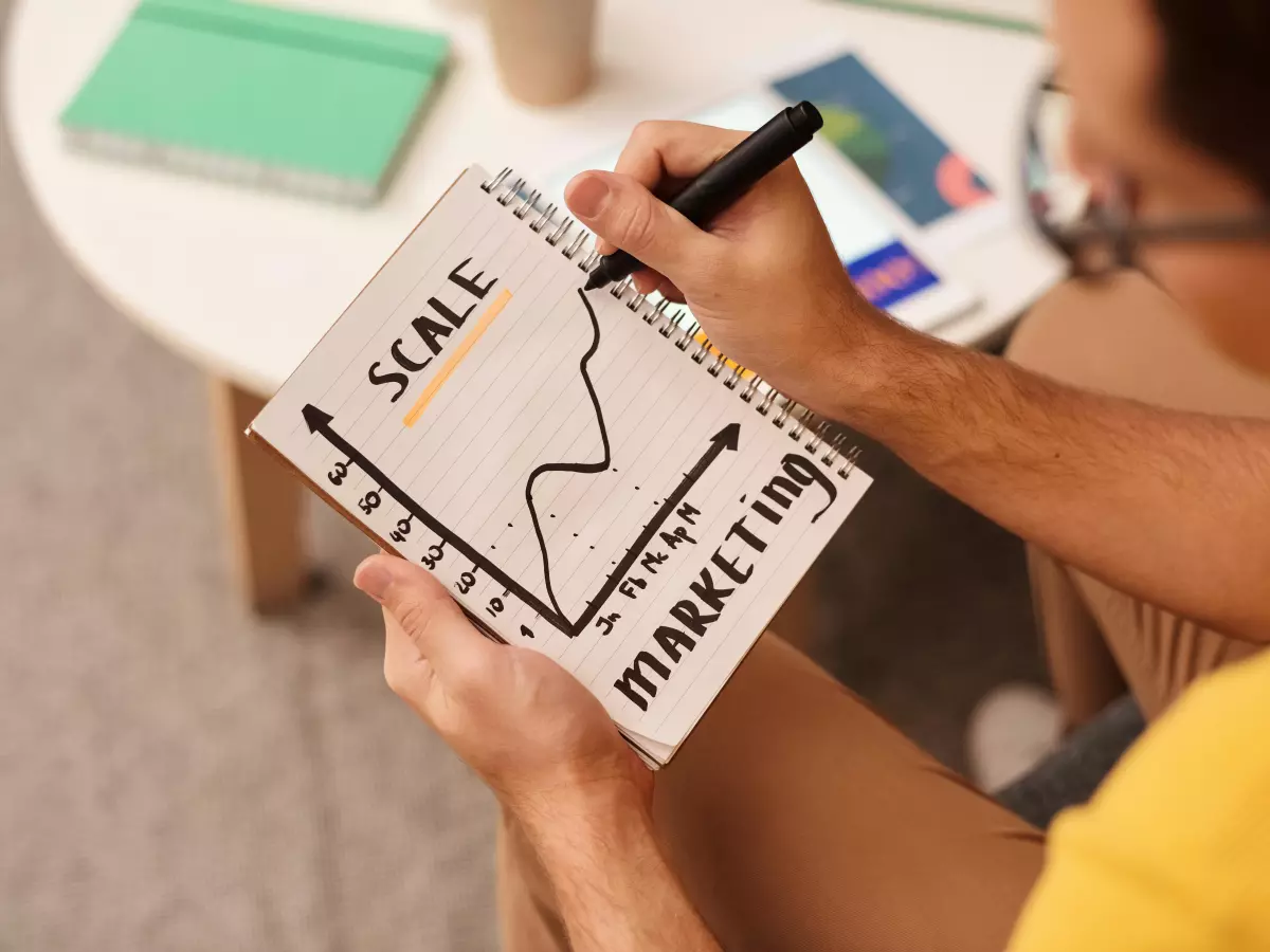 A person in a yellow shirt is writing "SCALE" on a notebook with a graph related to market growth, illustrating the importance of scaling in machine learning.