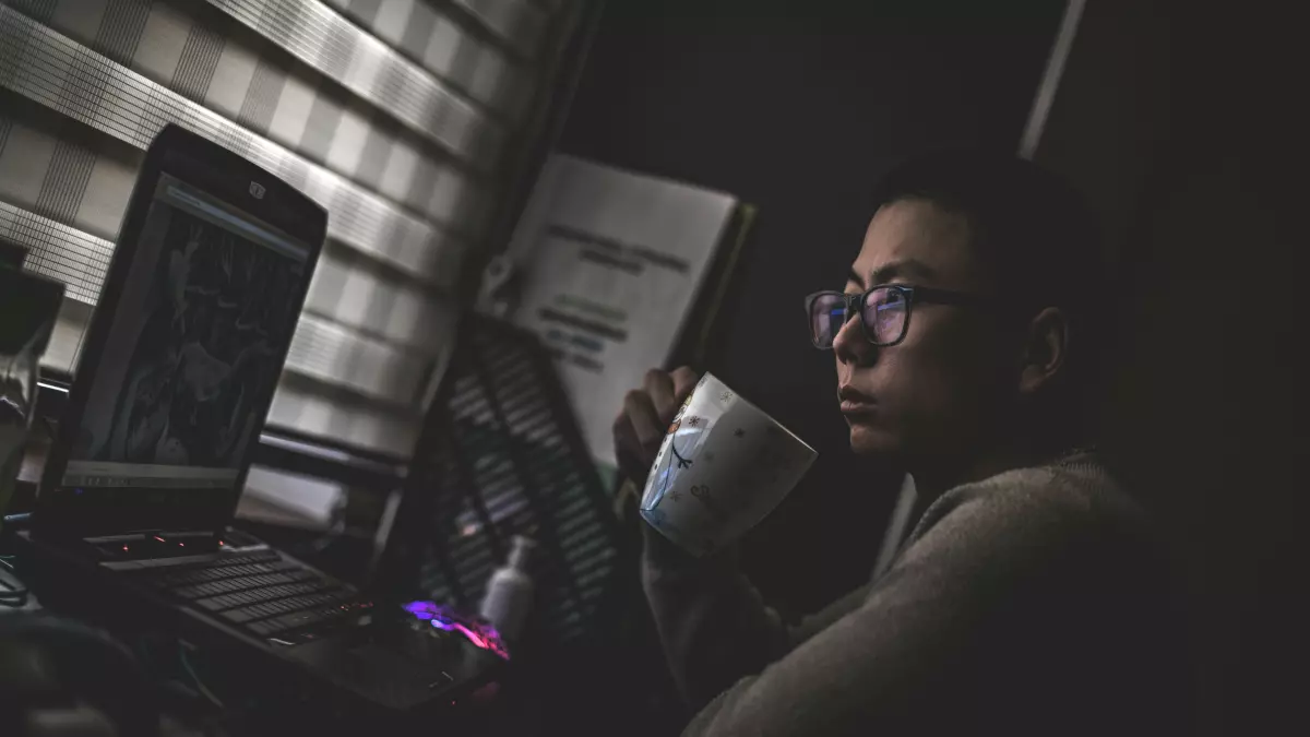 A man in a grey sweater is sitting at a desk in front of a laptop, looking at the screen. He is holding a cup in his right hand.  He is wearing glasses, and the blinds behind him are drawn.