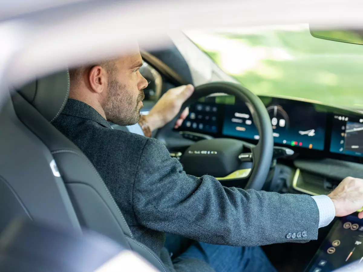 A man in a gray suit driving a car, focused on the road. You can see the car's interior with a steering wheel, dashboard and a big touchscreen.