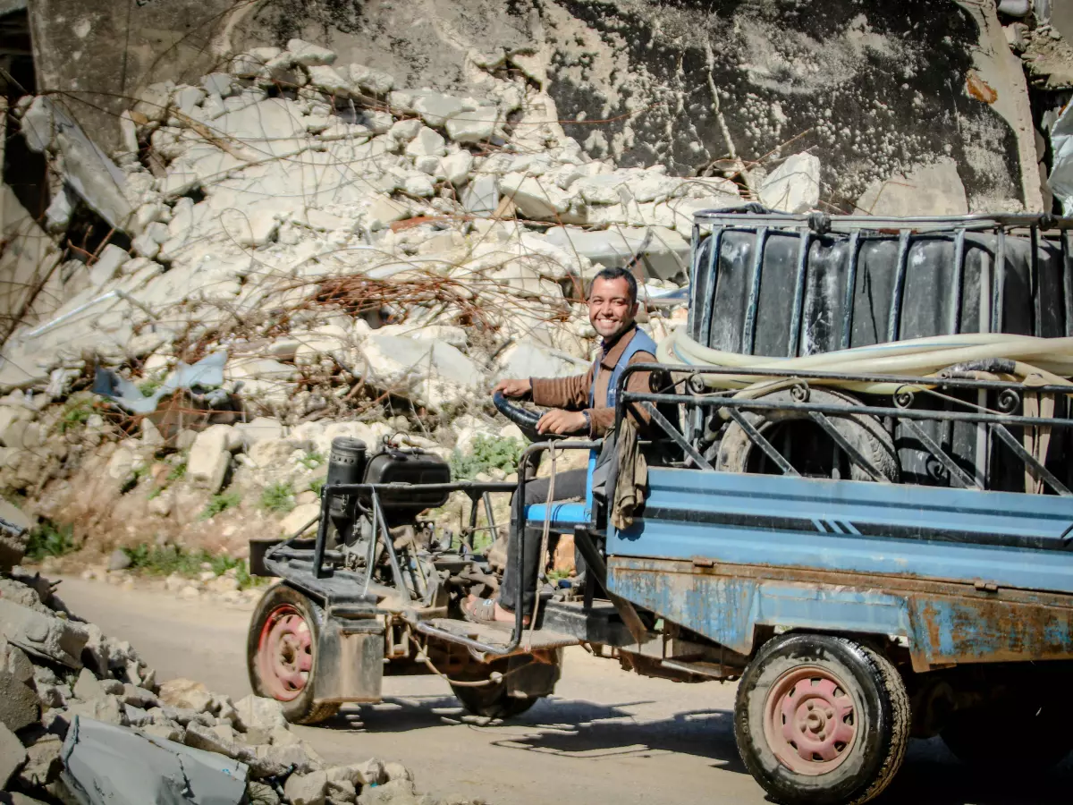 A man drives a blue vehicle past a ruined building. The building is covered in debris and dust.