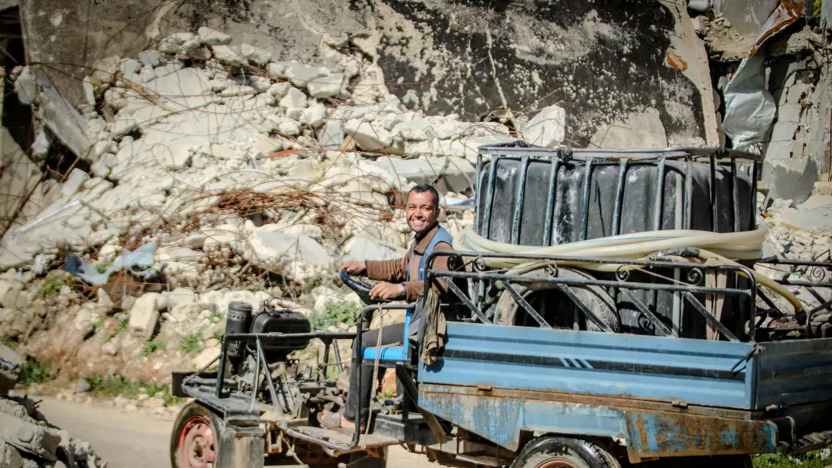 A man drives a blue vehicle past a ruined building. The building is covered in debris and dust.