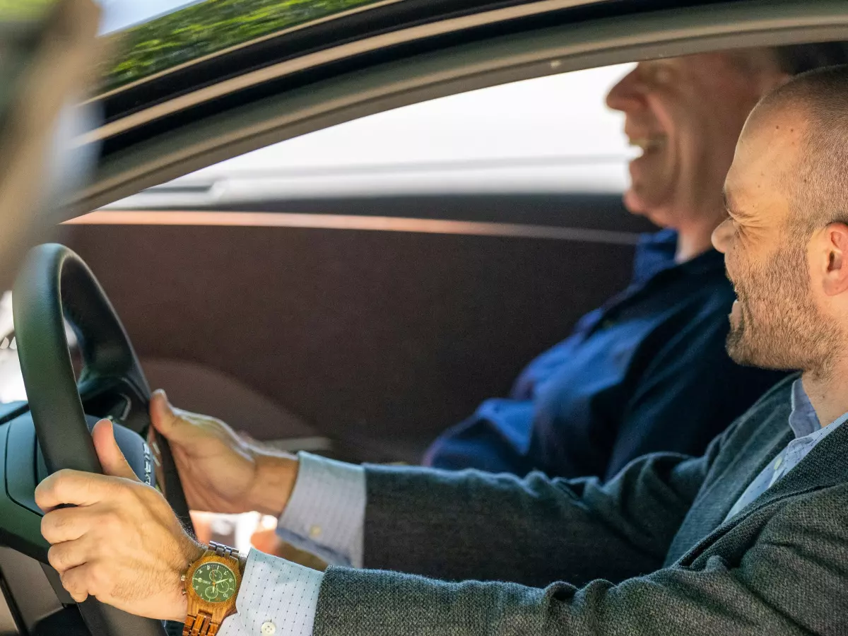 A man in a suit is driving a car and smiling, with a younger man in the passenger seat.