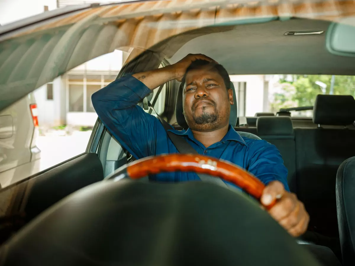 A man in a blue shirt is sitting in a car with his hands on his head, looking frustrated.