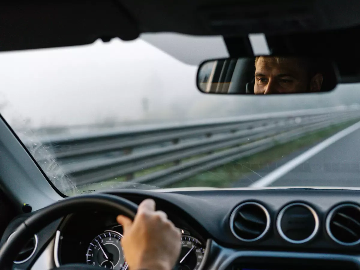 A driver's hand on a steering wheel, with a blurry view of the road ahead and the driver's reflection in the rearview mirror.