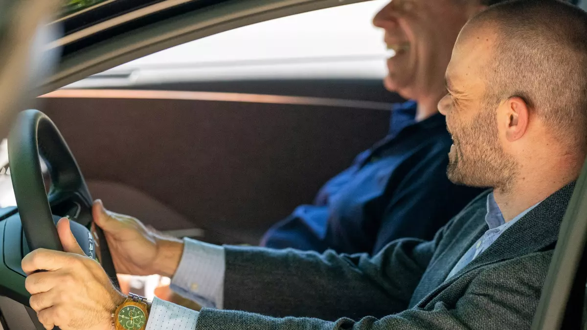 A man in a suit is driving a car and smiling, with a younger man in the passenger seat.