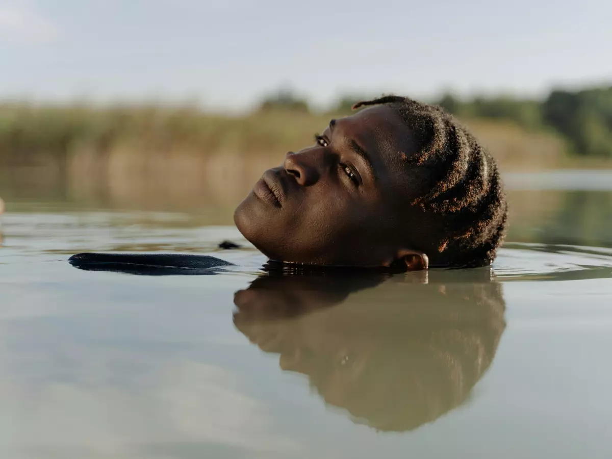 A person with long black hair is submerged in water, only their head and part of their shoulders are visible. The person is looking up at the sky. The water is dark and the sky is bright blue.