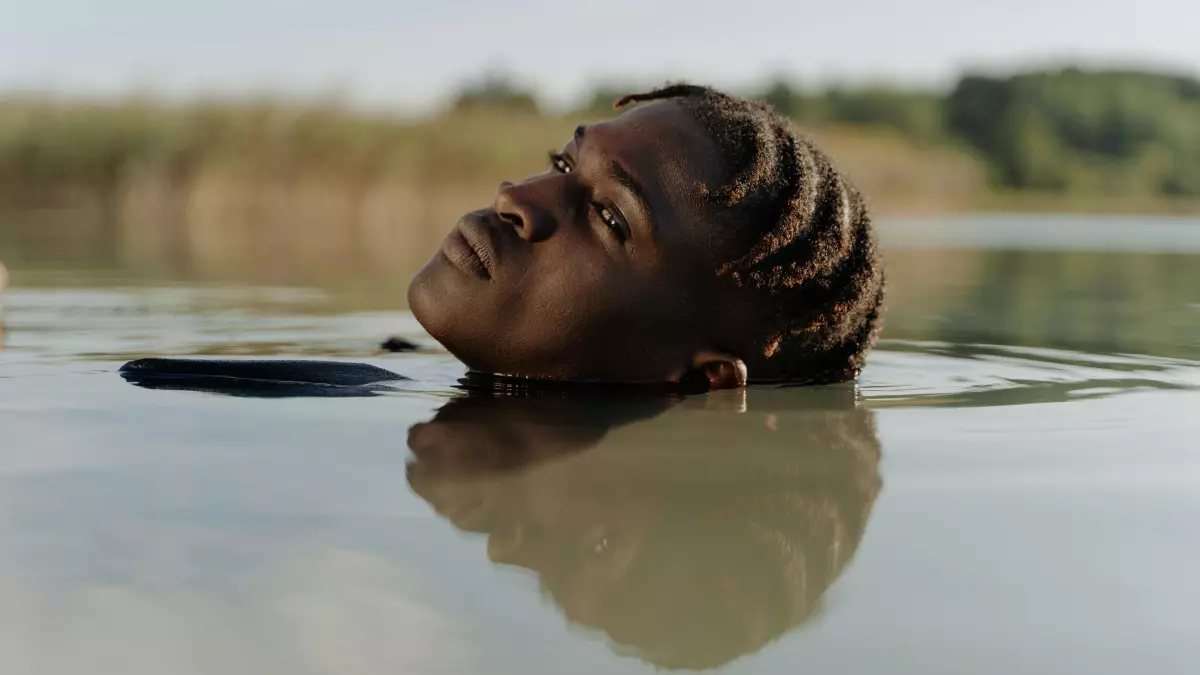 A person with long black hair is submerged in water, only their head and part of their shoulders are visible. The person is looking up at the sky. The water is dark and the sky is bright blue.