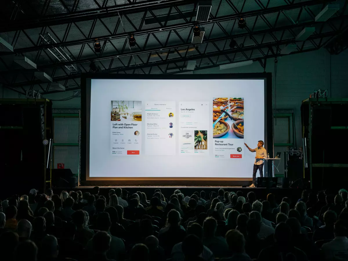 A man in a yellow shirt is standing on a stage in front of a large audience. The image is taken from a high angle, showing the man speaking and pointing at a screen with multiple visuals. The audience is sitting in rows of chairs, focused on the man on the stage.