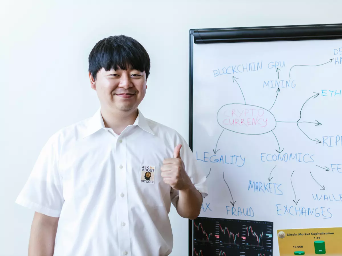 A young man with short black hair standing in front of a whiteboard with a diagram drawn on it. He is smiling and pointing to the board with his right hand.