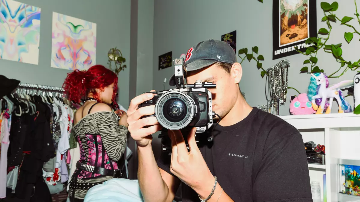 A man is holding a camera in his hands, he is looking at the lens and his focus is on the camera, behind him there is a woman in the background