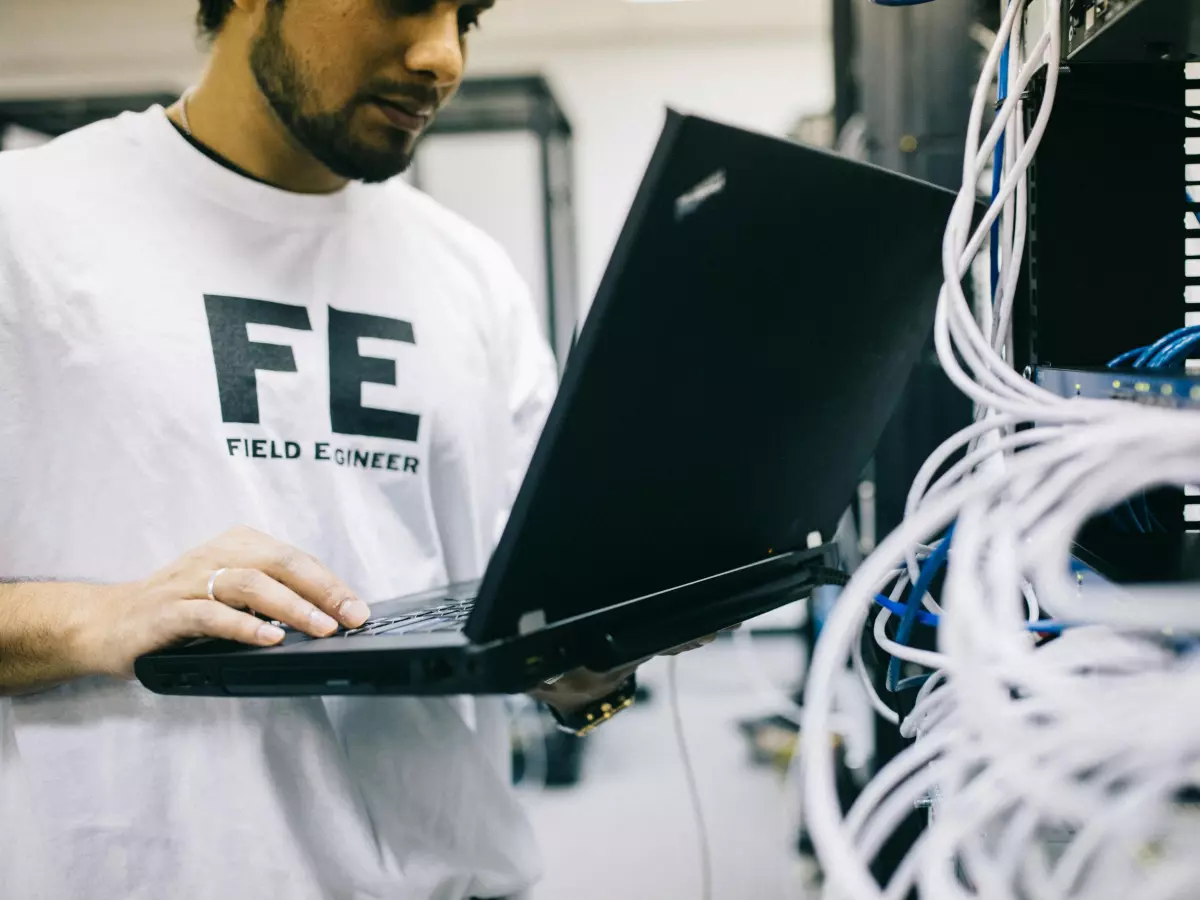 A man in a white t-shirt works on a laptop in a server room, surrounded by wires and cables.