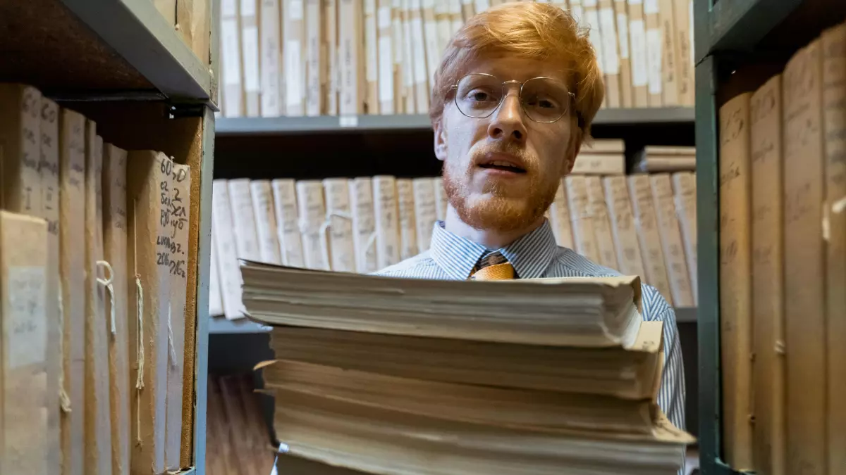 A man in glasses wearing a white shirt and a tie, holding a stack of books in a library setting. The books are filled with text, creating a visual representation of data. 