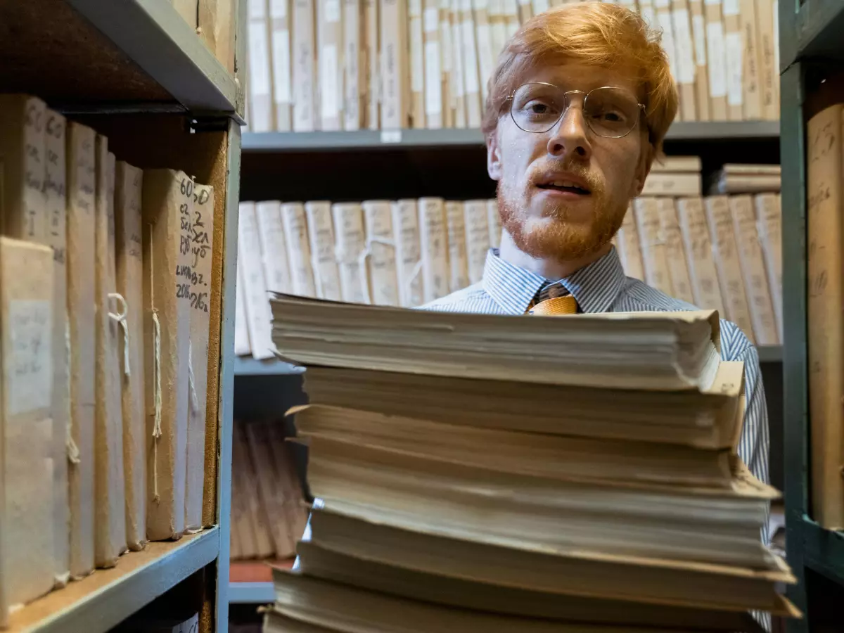 A man in glasses wearing a white shirt and a tie, holding a stack of books in a library setting. The books are filled with text, creating a visual representation of data. 