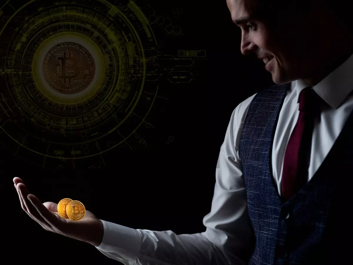 A man in a suit is holding two Bitcoin coins in his hand, with a large Bitcoin logo behind him.