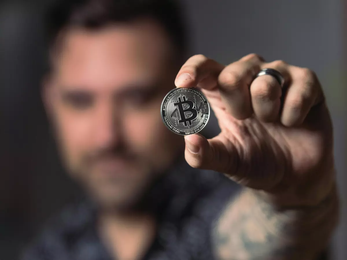 A man with a tattoo holds a Bitcoin coin in front of a blurry background.