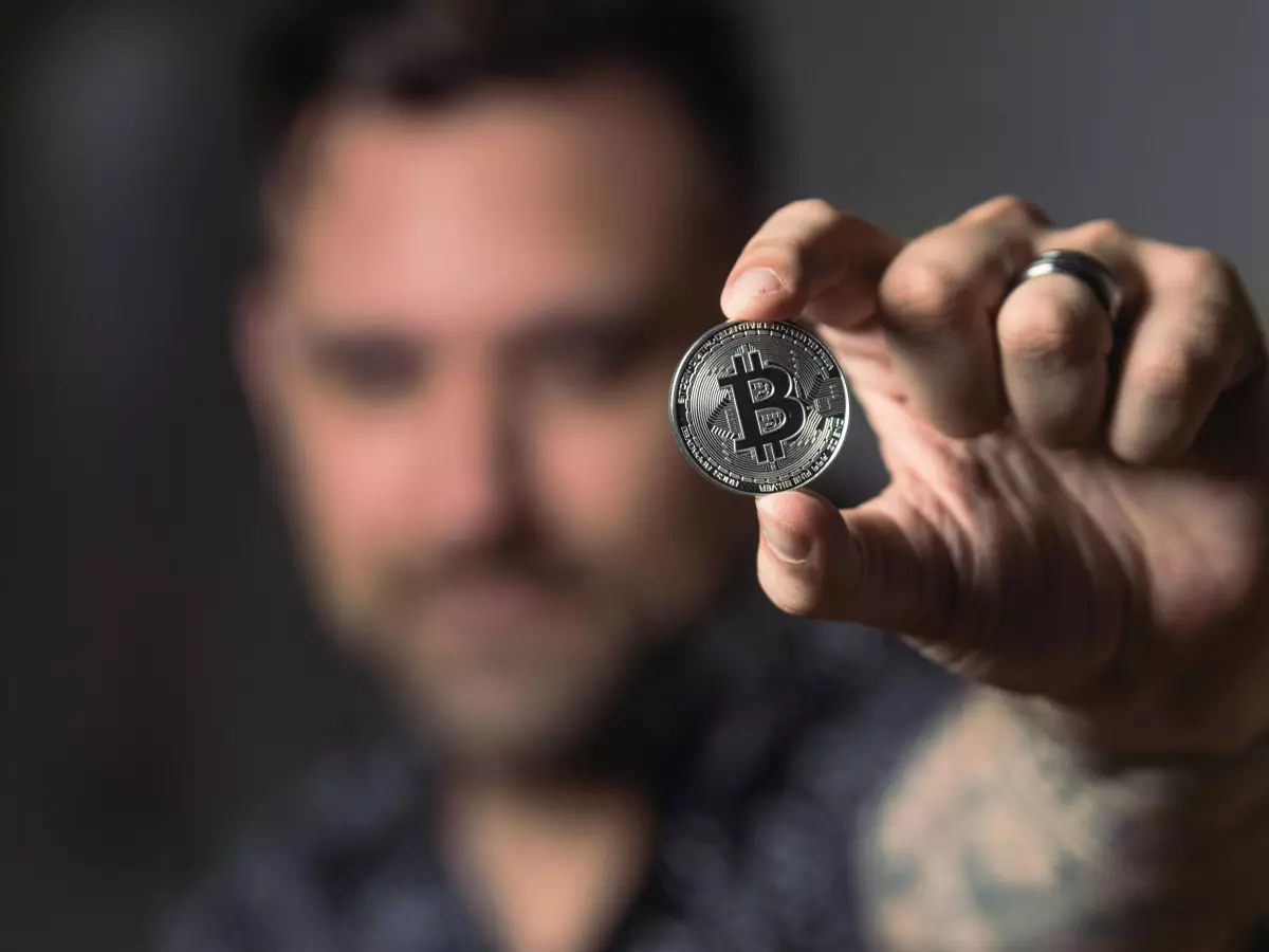 A close-up shot of a person's hand holding a Bitcoin coin, with the person's face blurred in the background.