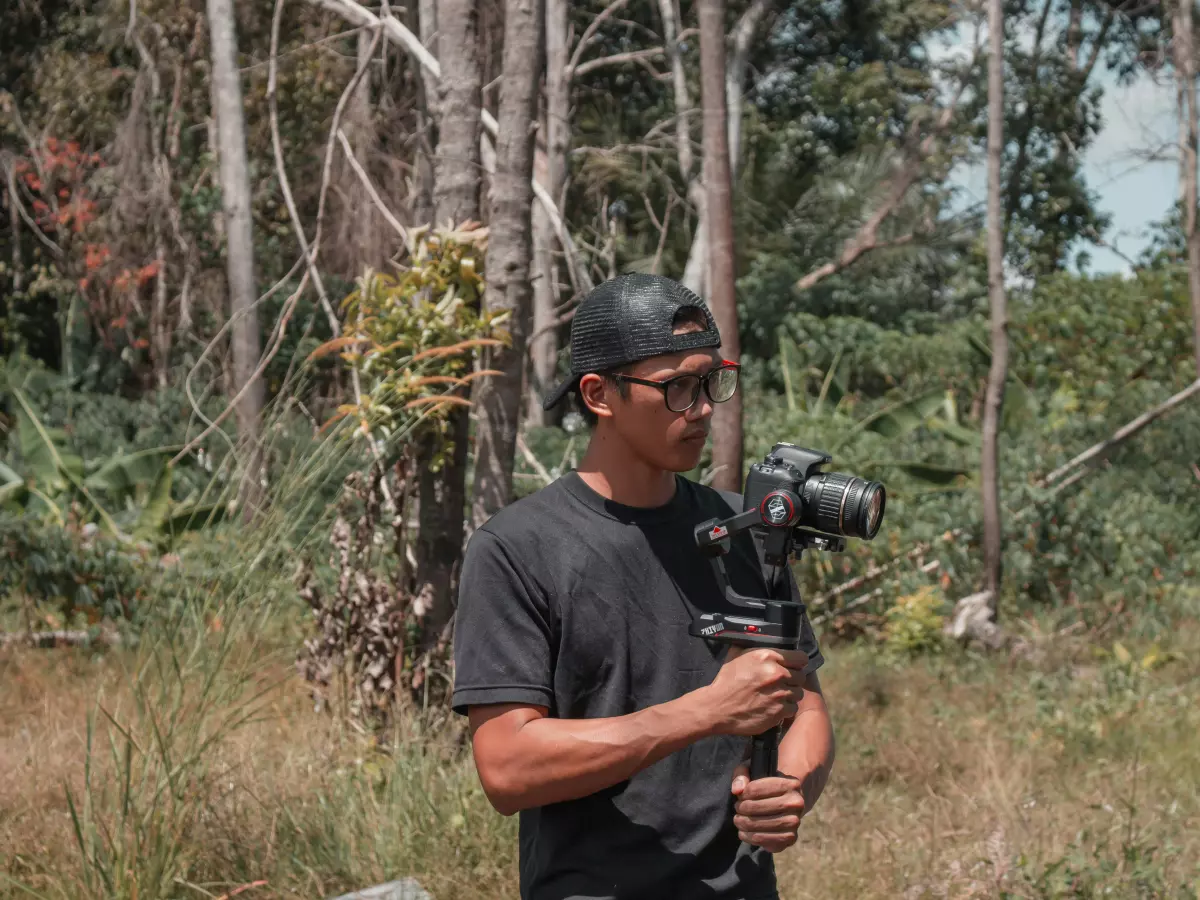 A man is holding a camera on a tripod in a forest setting. He is wearing a black shirt and a cap, and is looking at the camera.