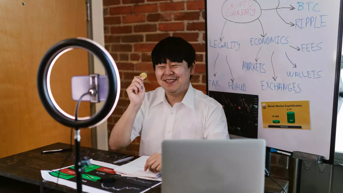 A young man is sitting at a desk in front of a laptop. He is wearing a white shirt and has short black hair. He is smiling and holding a small object in his right hand. There is a ring light behind him, and a smartphone is attached to it. There are also some papers and a book on the desk. The background is a brick wall.