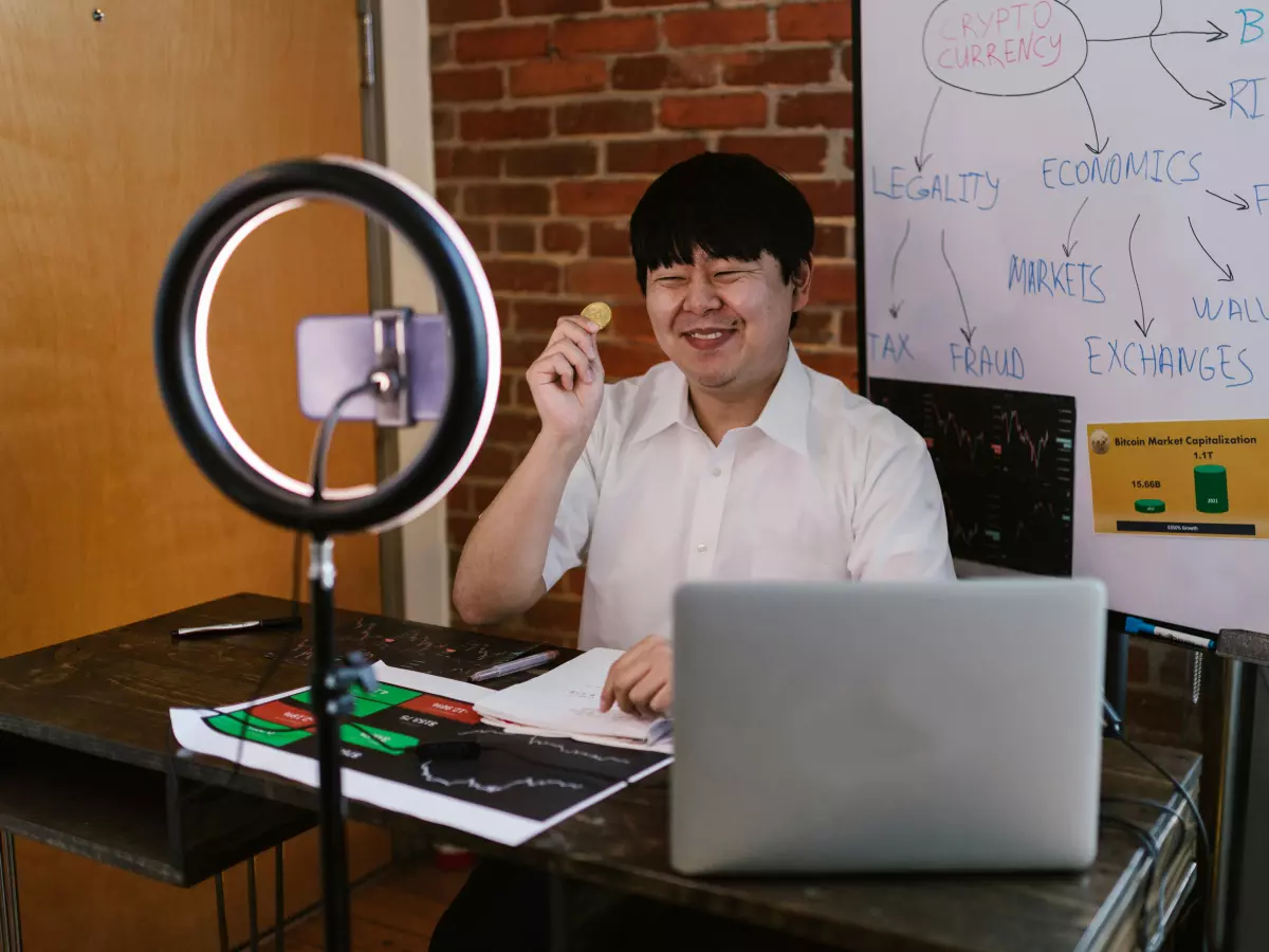 A young man is sitting at a desk in front of a laptop. He is wearing a white shirt and has short black hair. He is smiling and holding a small object in his right hand. There is a ring light behind him, and a smartphone is attached to it. There are also some papers and a book on the desk. The background is a brick wall.