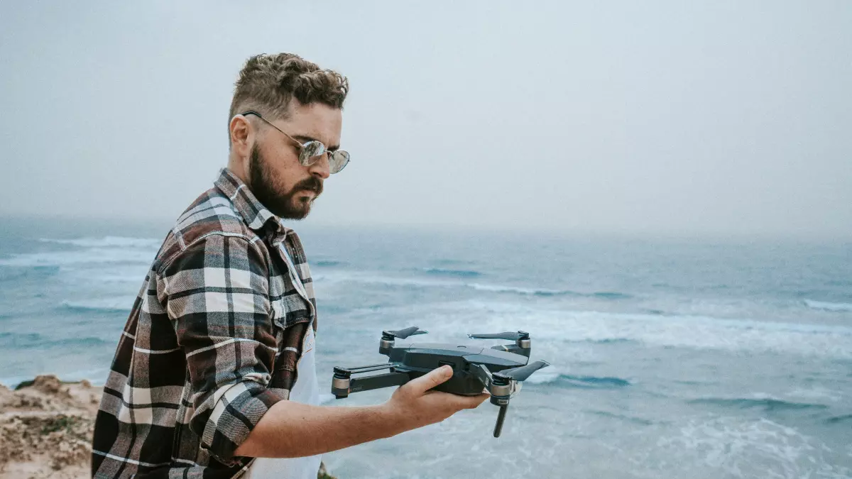 A man in a plaid shirt and sunglasses is standing on a cliff overlooking the ocean, holding a small black drone in his hands. 