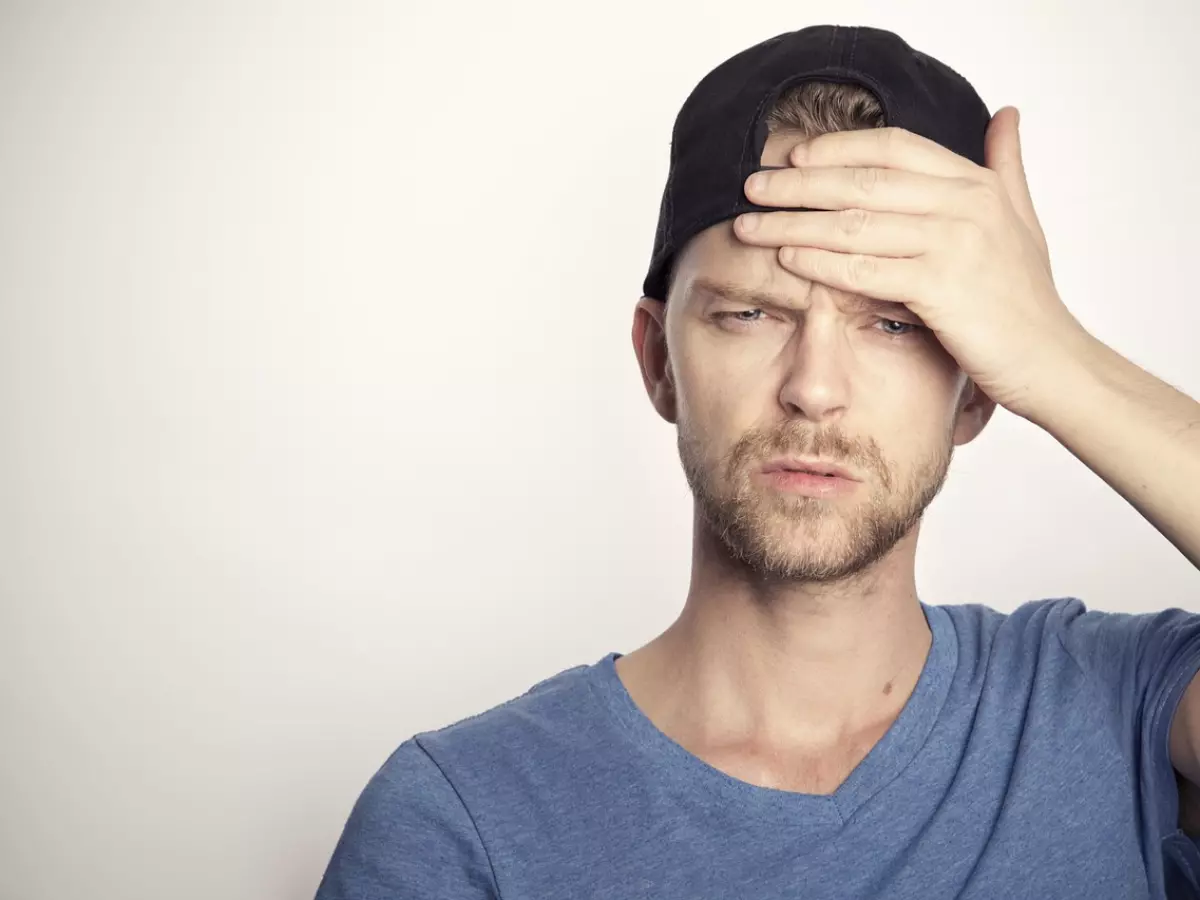 A man in a blue shirt and a black cap is standing in front of a white background, looking distressed, one hand is on his forehead.  The image captures a look of intense frustration.