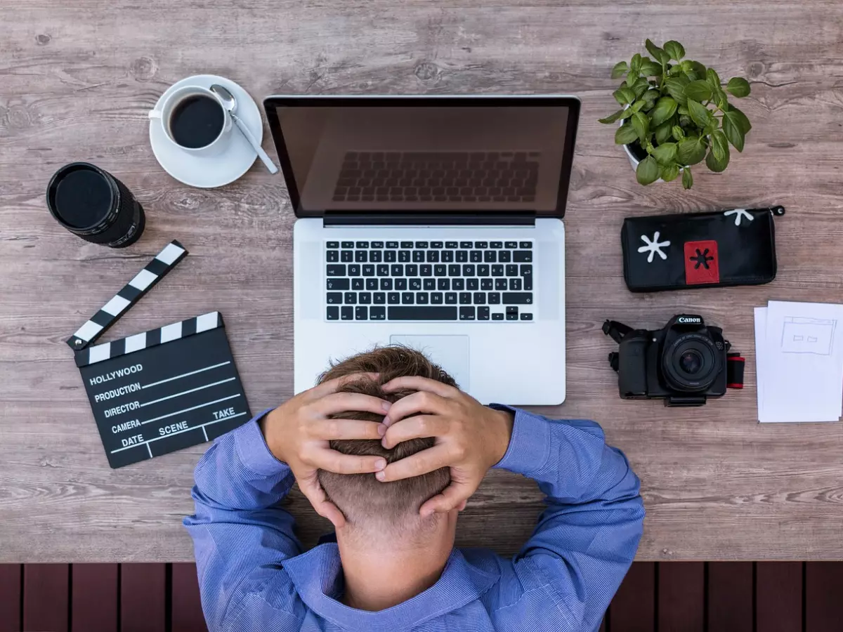 A man sits at a desk with his head in his hands, looking stressed. There is a laptop, a camera, a coffee cup, and other items on the desk. 