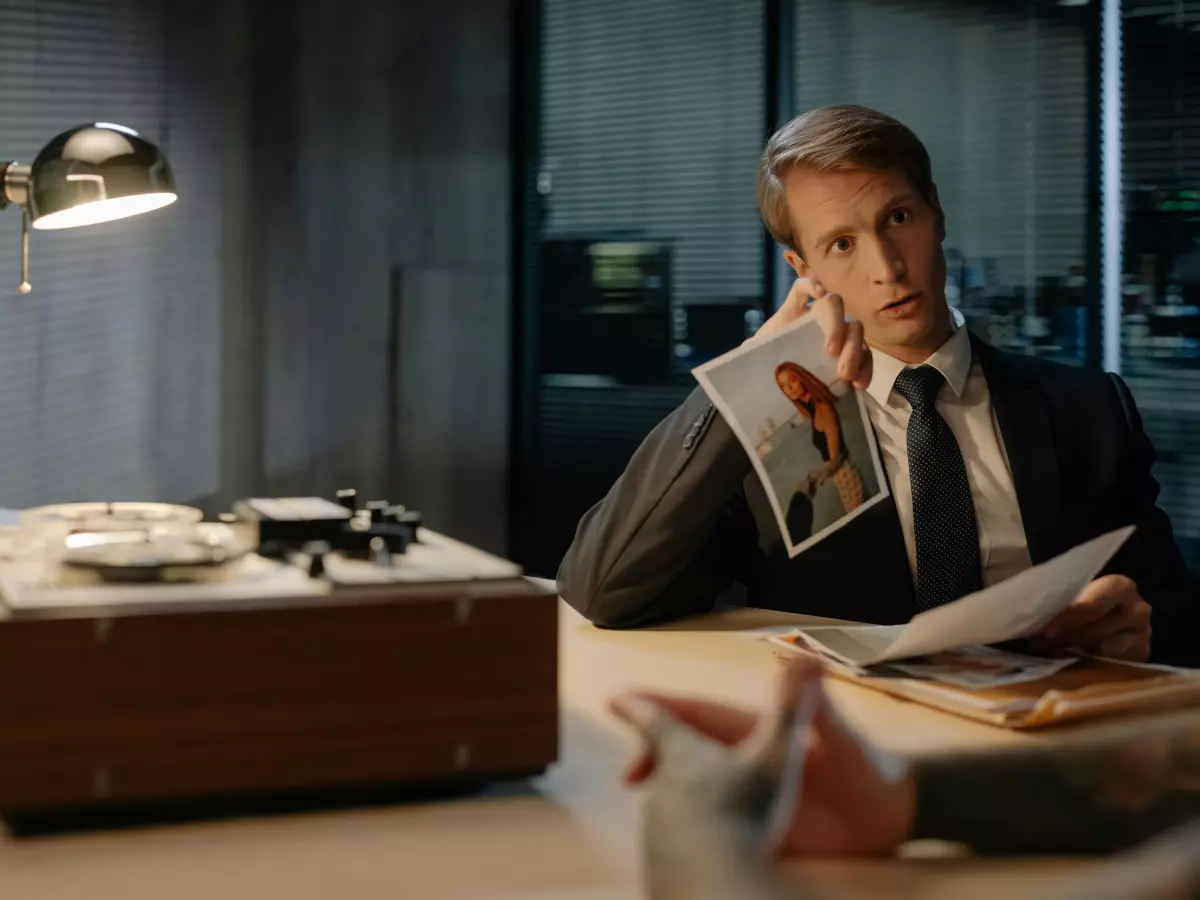 A man in a suit sits at a table in a room with a gray wall, holding a paper in his hand.  He looks directly into the camera, with a serious and focused expression. 