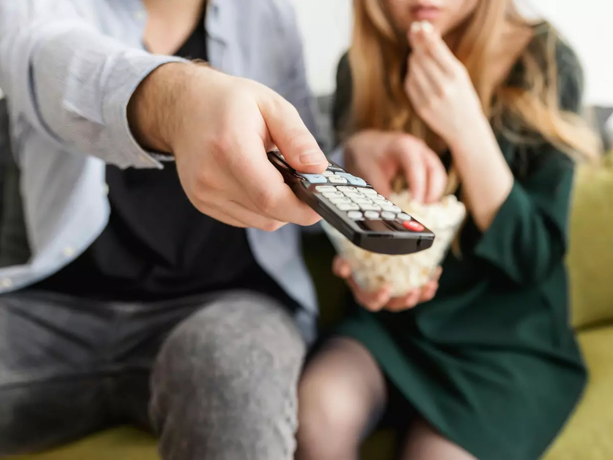 Two people are watching TV on a couch. One person is using the remote control.