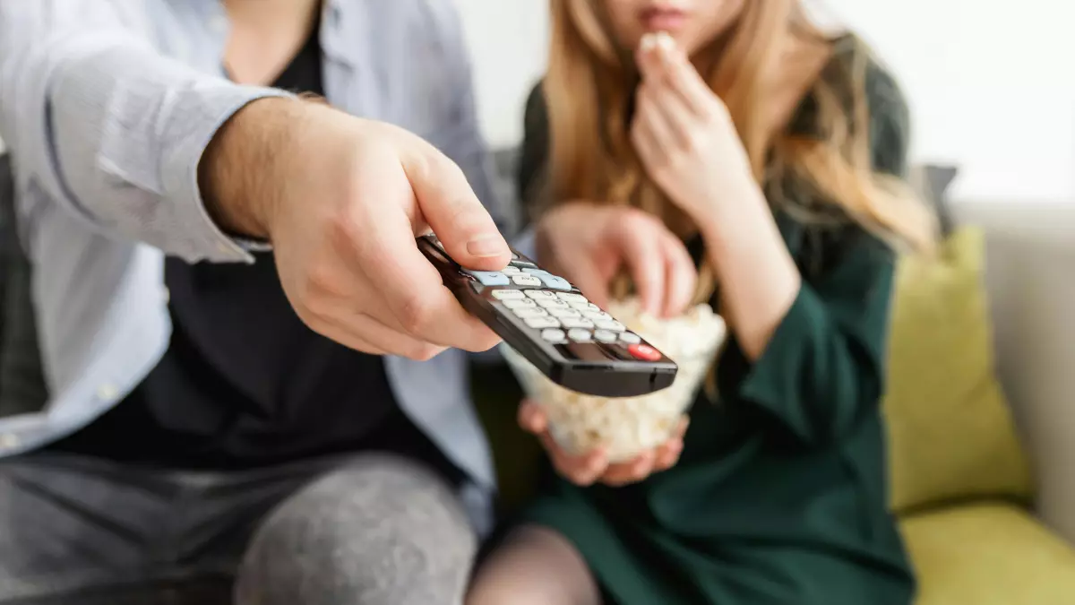 Two people are watching TV on a couch. One person is using the remote control.