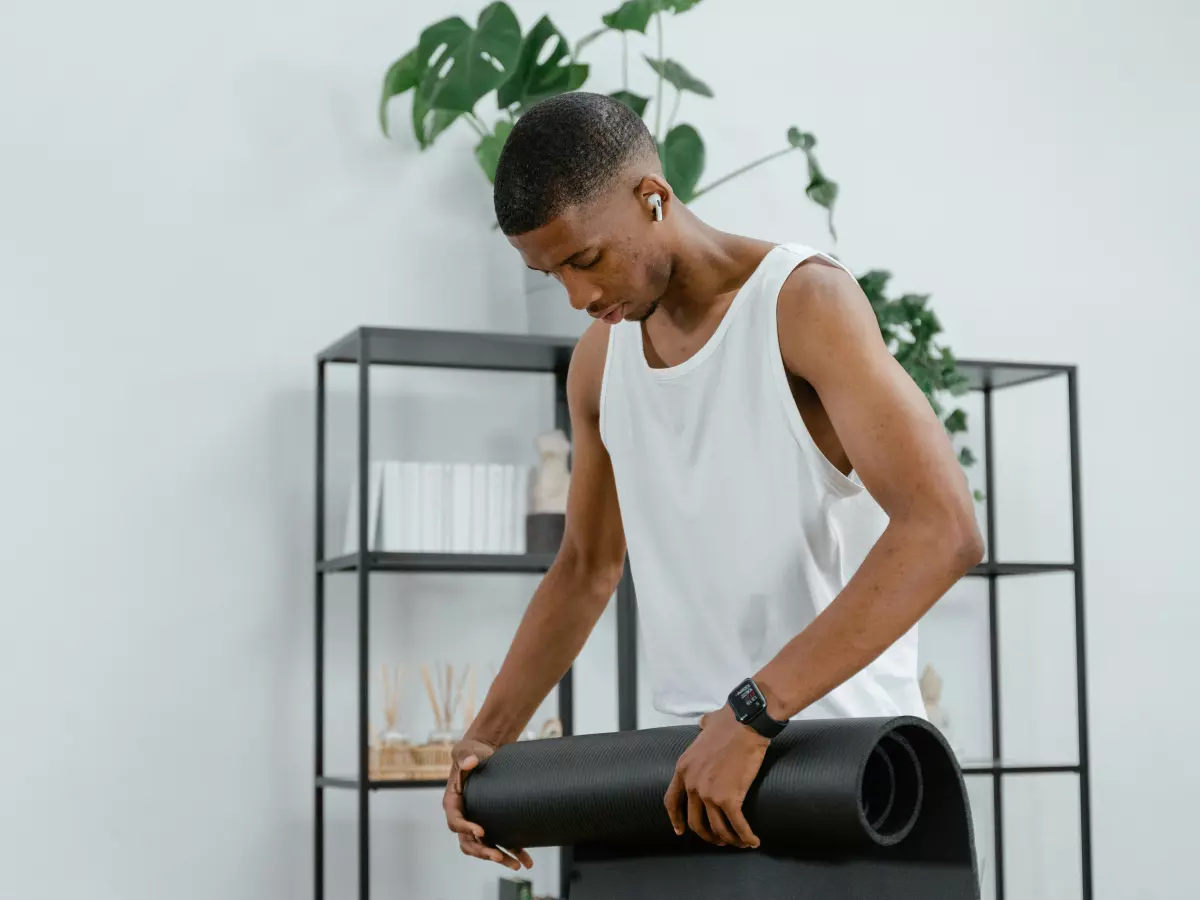 A man wearing a smartwatch and a white tank top rolls up a black yoga mat. He is looking at the mat as he rolls it up. He is standing in front of a white wall with a green plant in a white pot on a shelf behind him.