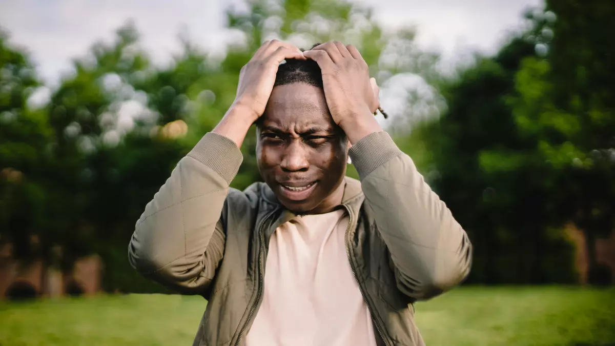 A person sitting on a grass field, with a laptop on their lap, looking at the laptop, covering their face with their hands, looking distressed.