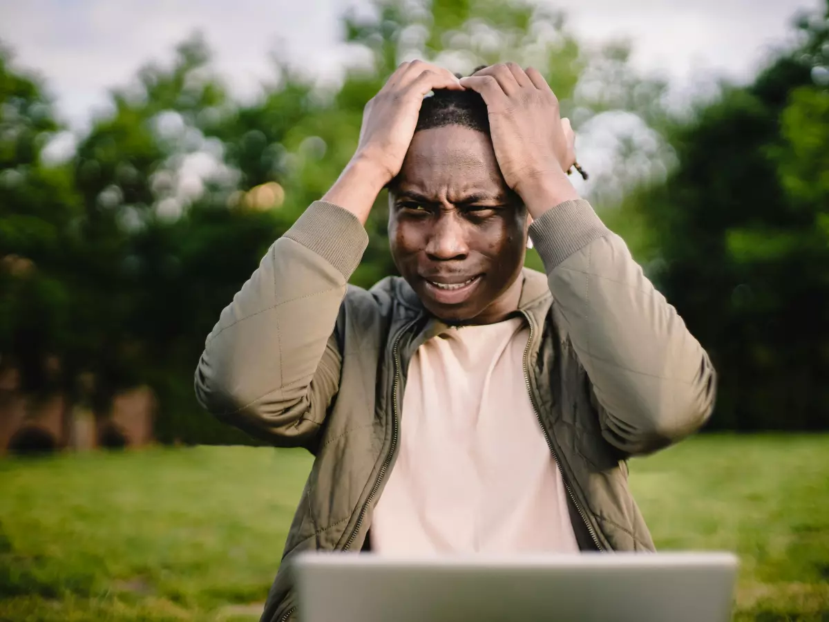 A person sitting on a grass field, with a laptop on their lap, looking at the laptop, covering their face with their hands, looking distressed.