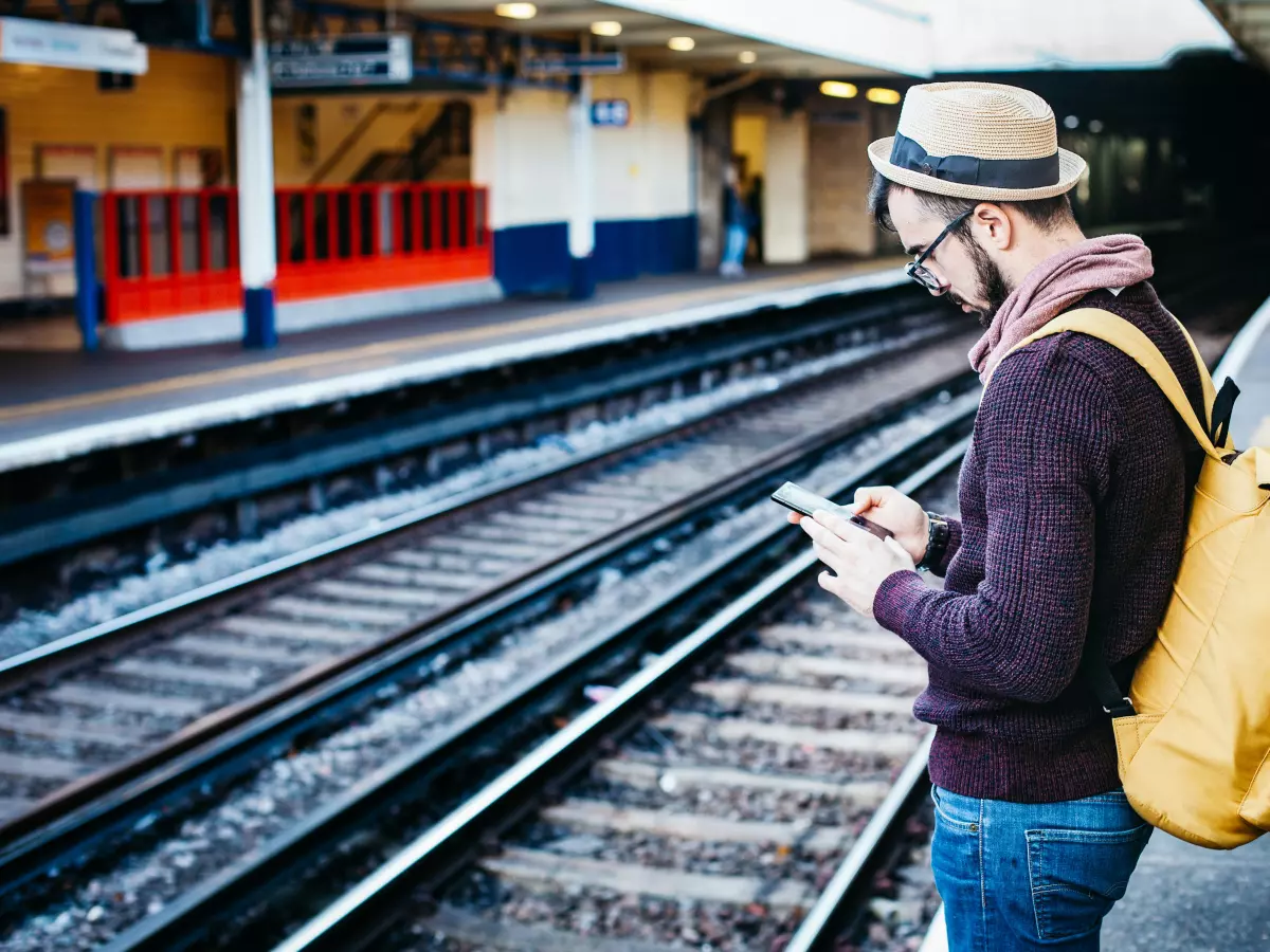 A young man is standing on a train platform, looking at his smartphone, with a yellow backpack on his back.