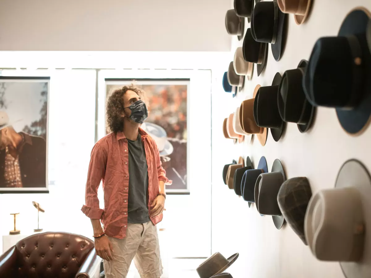 A young man is in a store, looking closely at something he's holding in his hand. His face is obscured by a mask, adding a sense of mystery and intrigue. He is surrounded by various items, suggesting a shopping environment.