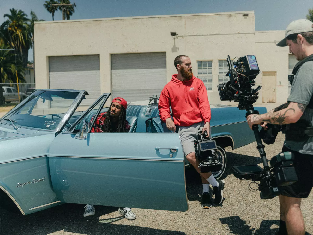 A film crew is shooting a scene with an actor in a blue convertible. The camera is on a gimbal and the cameraman is wearing a harness. The actor is wearing a red hoodie and bandana. The background is a warehouse.