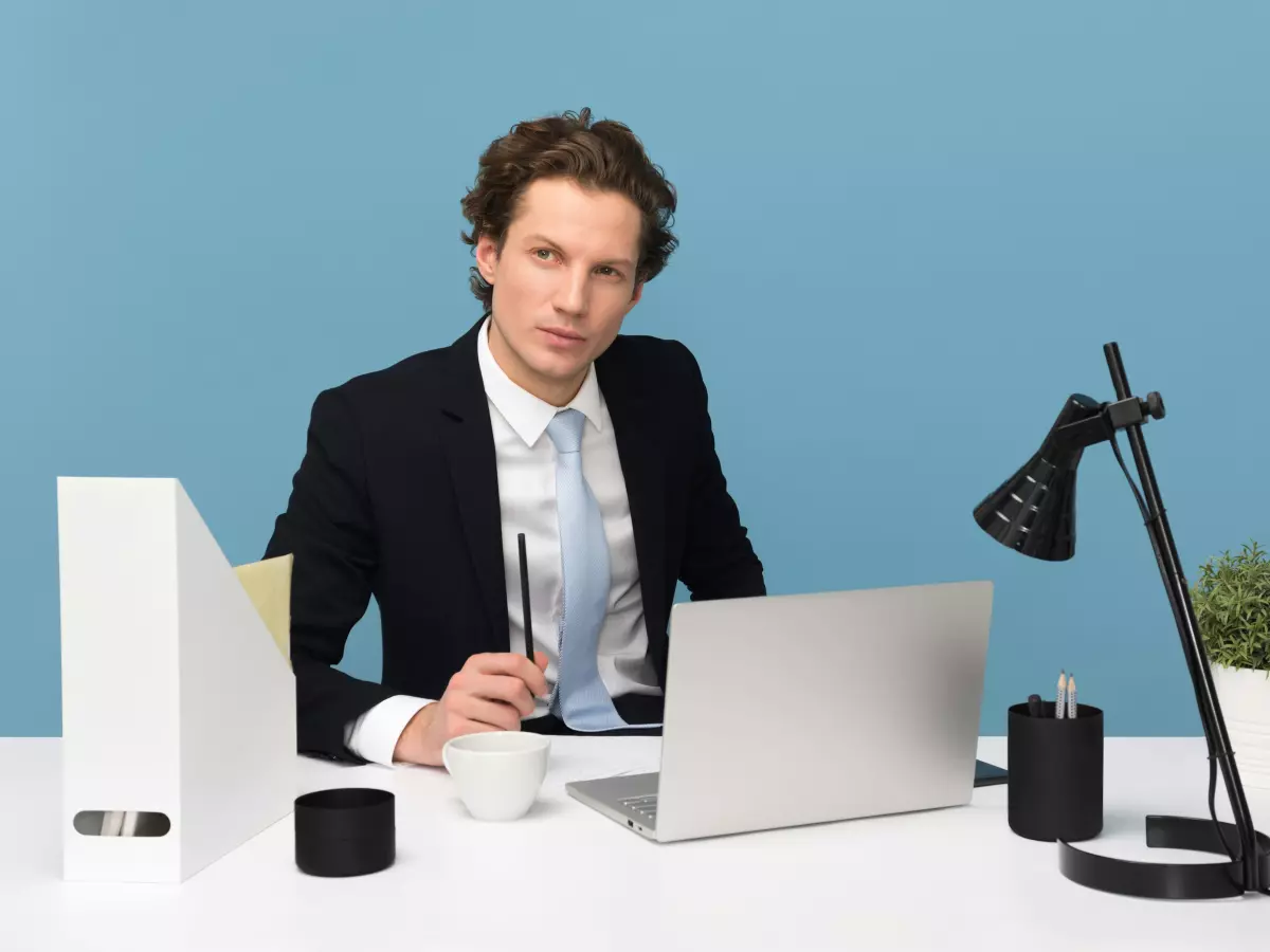 A man in a suit sits at a desk in front of a laptop. He is looking at the camera with a serious expression on his face. There is a lamp, a plant, a cup of coffee and some folders on the desk.