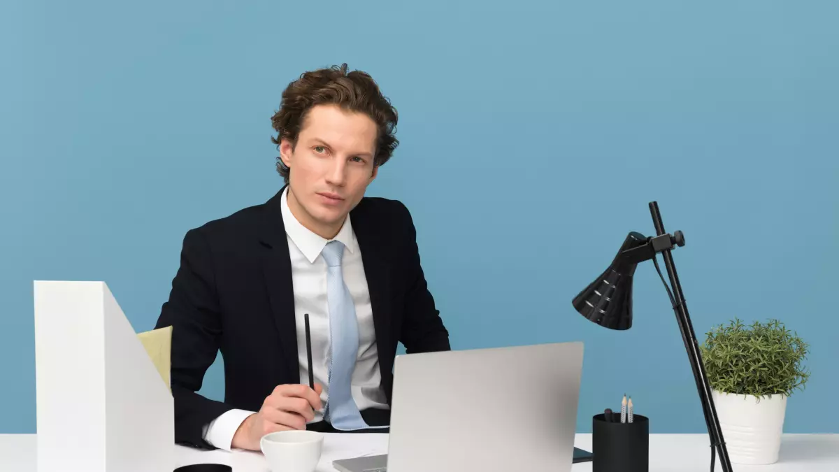 A man in a suit sits at a desk in front of a laptop. He is looking at the camera with a serious expression on his face. There is a lamp, a plant, a cup of coffee and some folders on the desk.