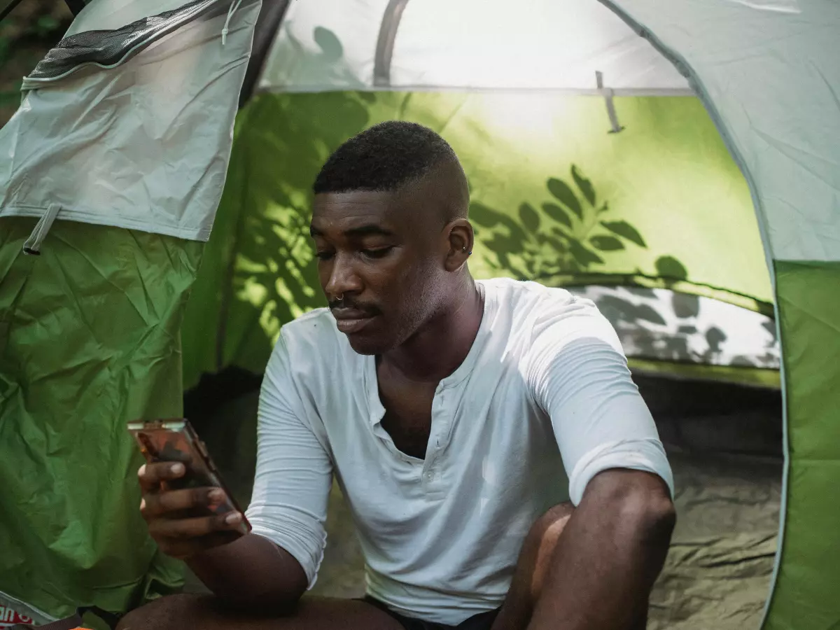 A young man sits inside a green tent, looking down at his phone. He is wearing a white shirt and has short black hair. The tent is surrounded by greenery.