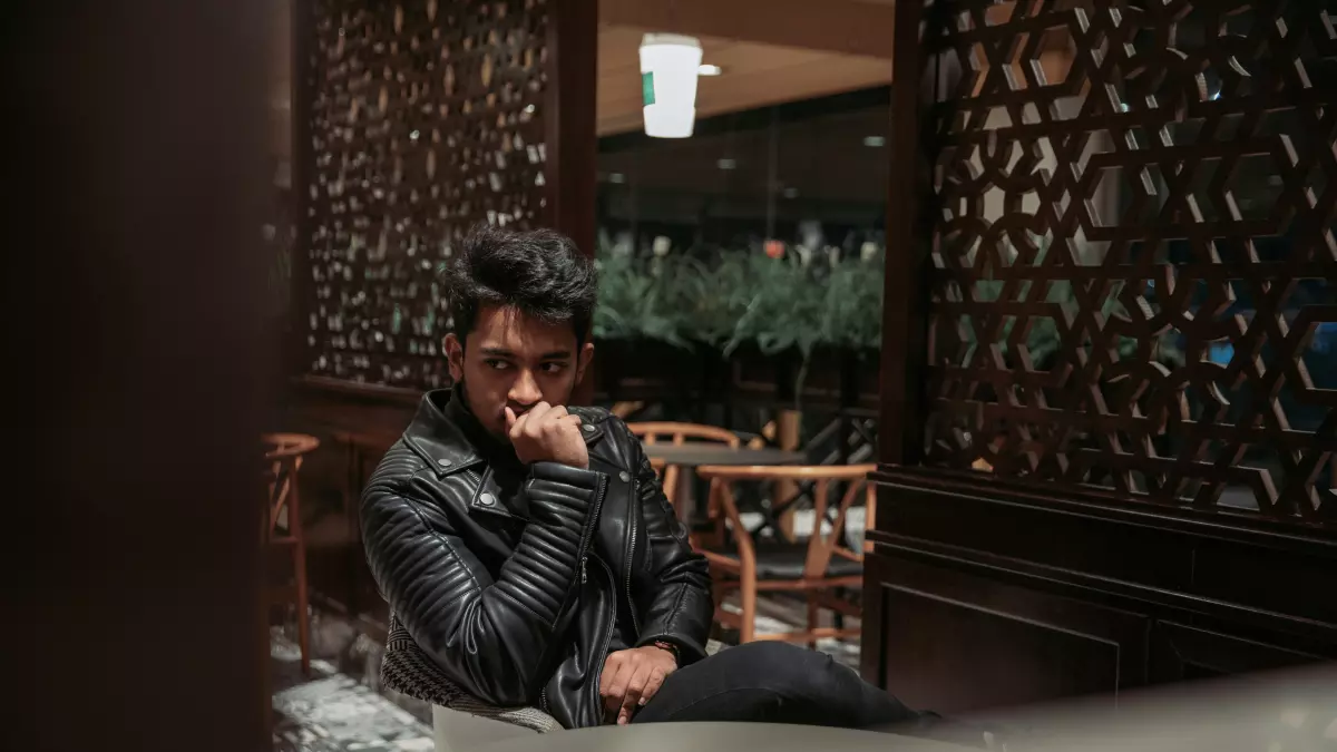 A man in a black leather jacket sits in a cafe, looking thoughtful.