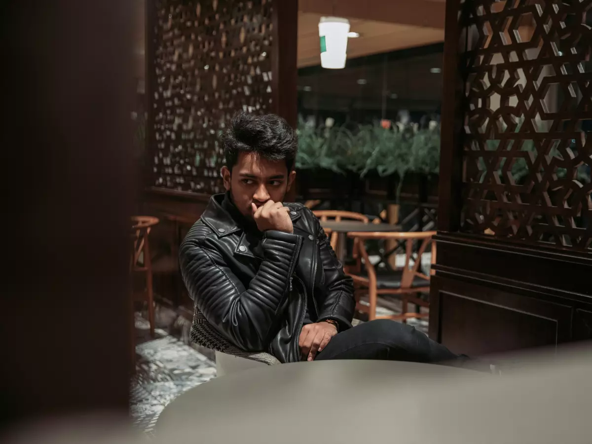 A man in a black leather jacket sits in a cafe, looking thoughtful.