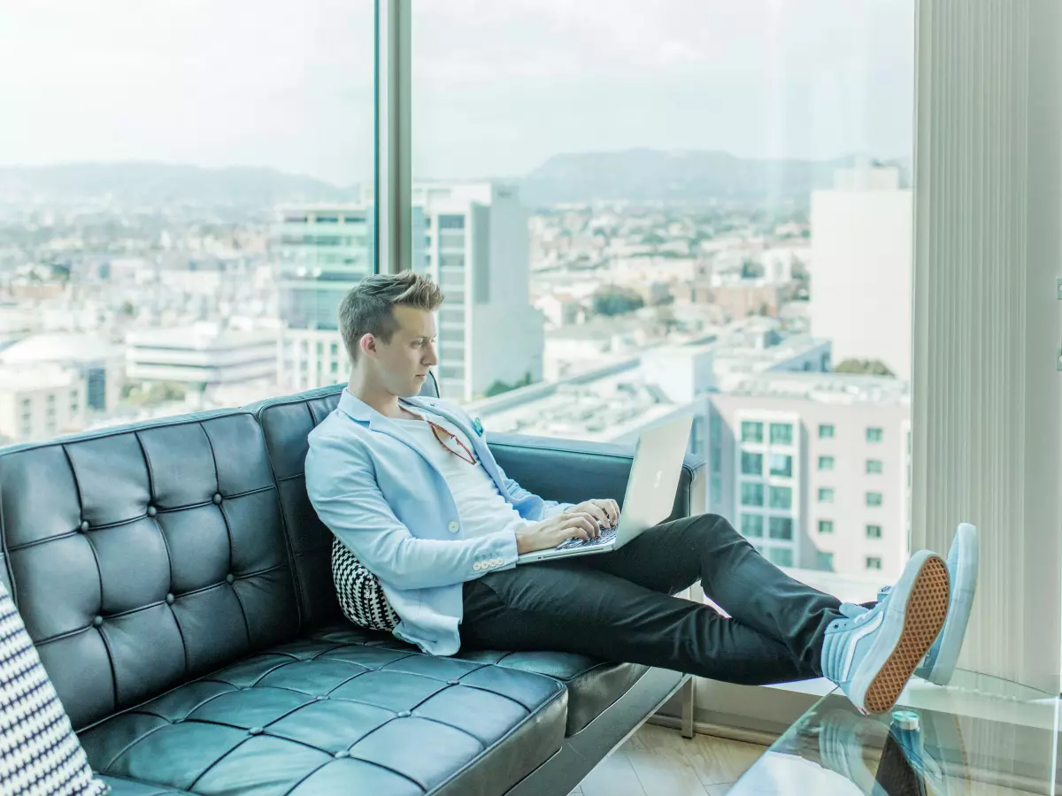 A man sitting on a couch, working on a laptop computer with a city skyline in the background.