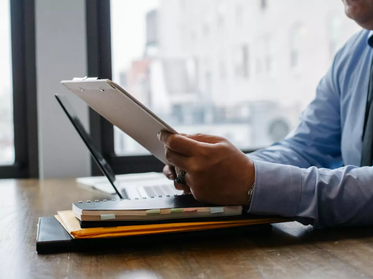 A person in a blue shirt and tie is sitting at a table looking at a tablet. A laptop and some folders are on the table. The person is wearing a business casual outfit.