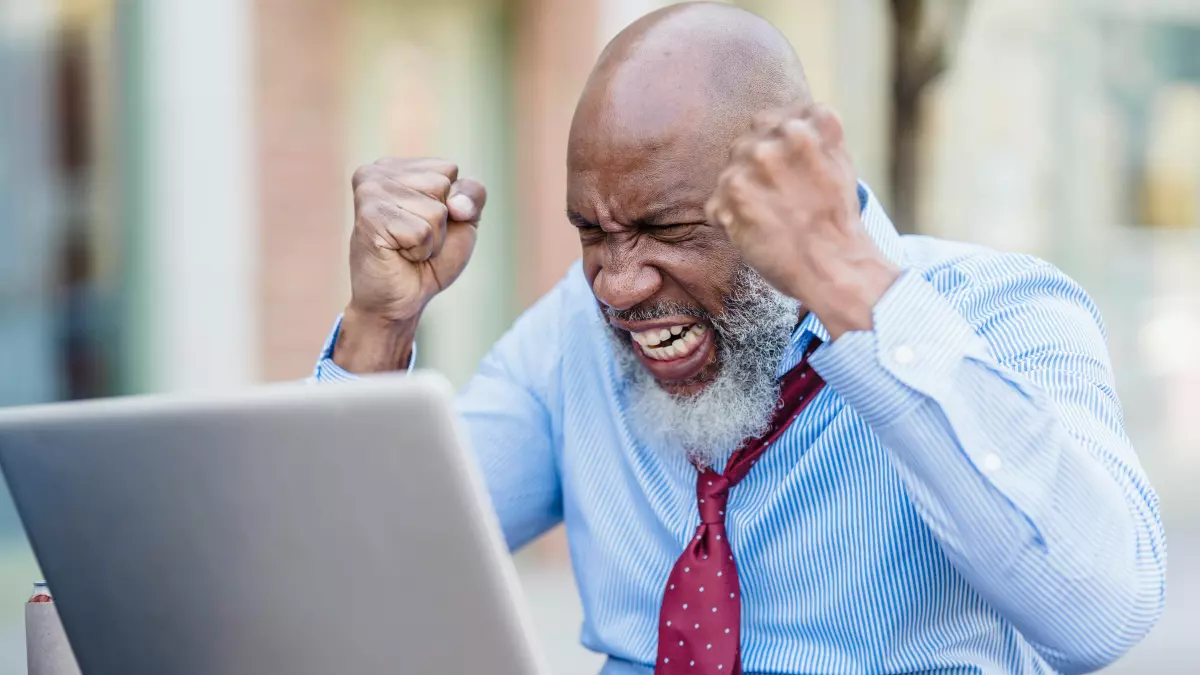 A black man in a blue shirt and red tie sitting on a bench with a laptop. He is holding his hands up in the air, as if he is frustrated.