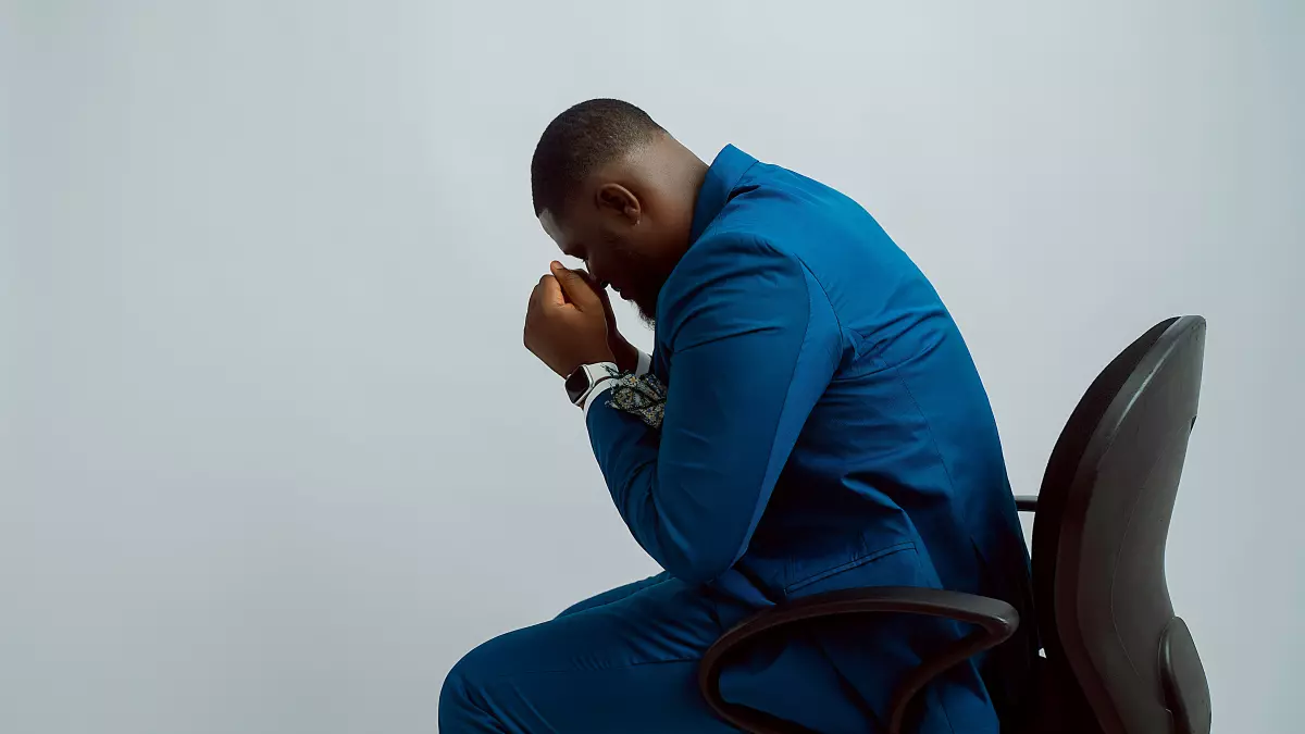 A man in a blue suit sits on a black office chair, his head in his hands. He appears to be in deep thought or distress.