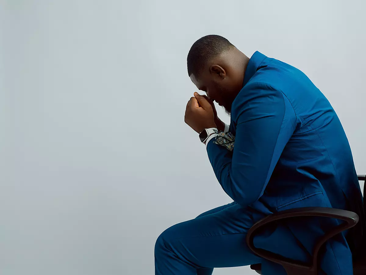 A man in a blue suit sits on a black office chair, his head in his hands. He appears to be in deep thought or distress.