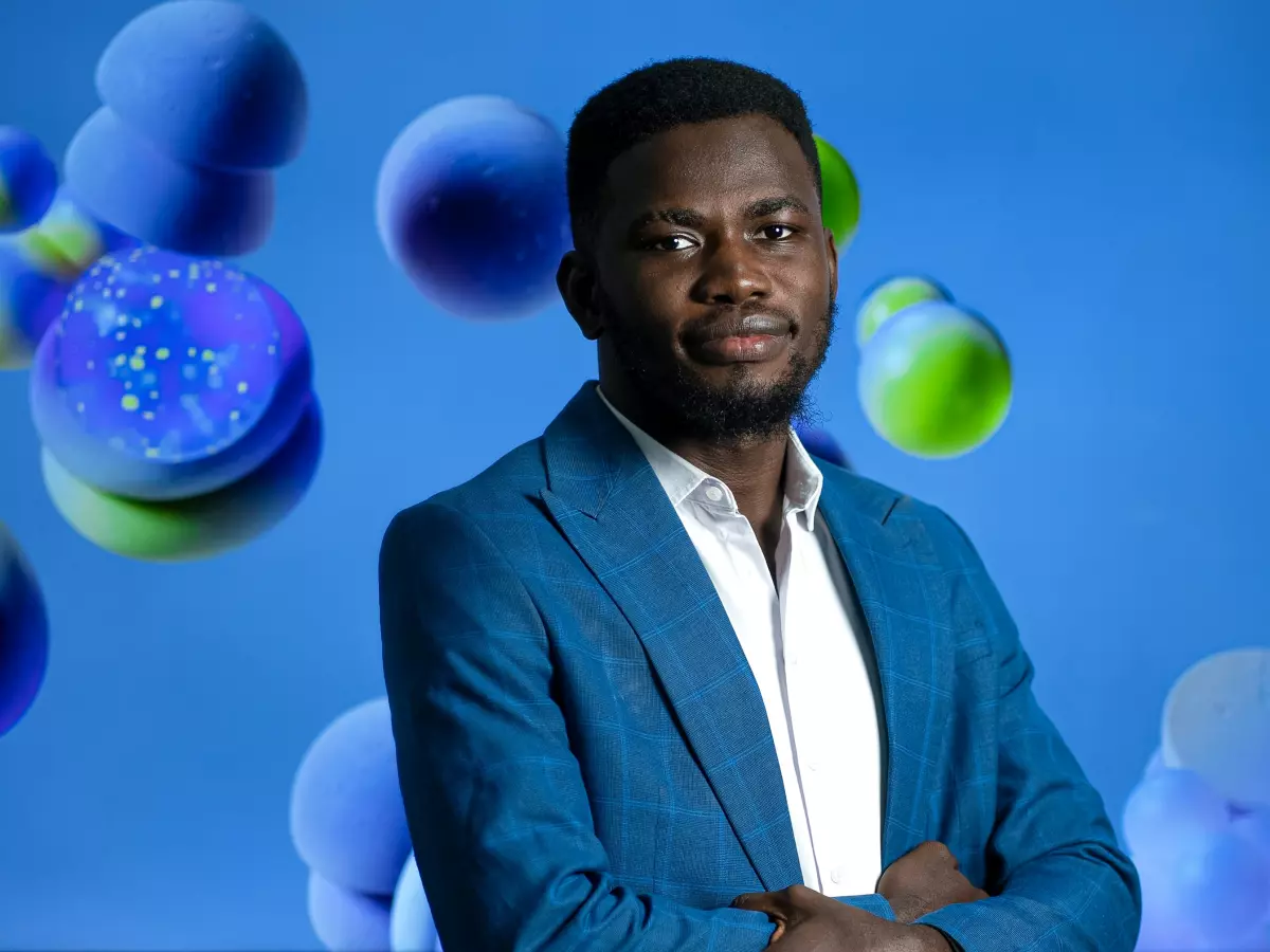 A young man with a beard and a blue blazer is standing in front of a blue background with abstract shapes. He is looking at the camera with a neutral expression and his arms are crossed. 