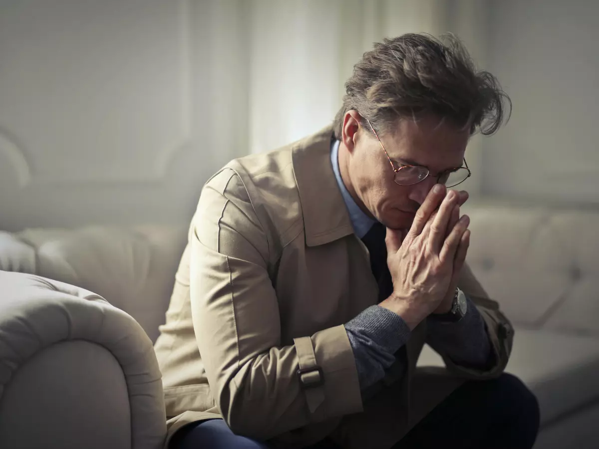 A man in a beige jacket sits on a sofa, his hands clasped together, with his head bowed and a pensive expression. The lighting is dim and the overall mood is somber.