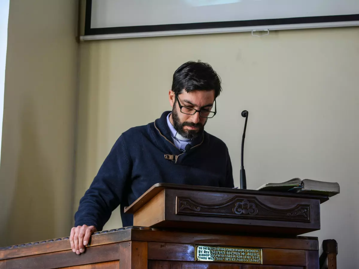 A man with a beard and glasses stands at a podium speaking into a microphone, a projection screen in the background, the scene is most likely in a church