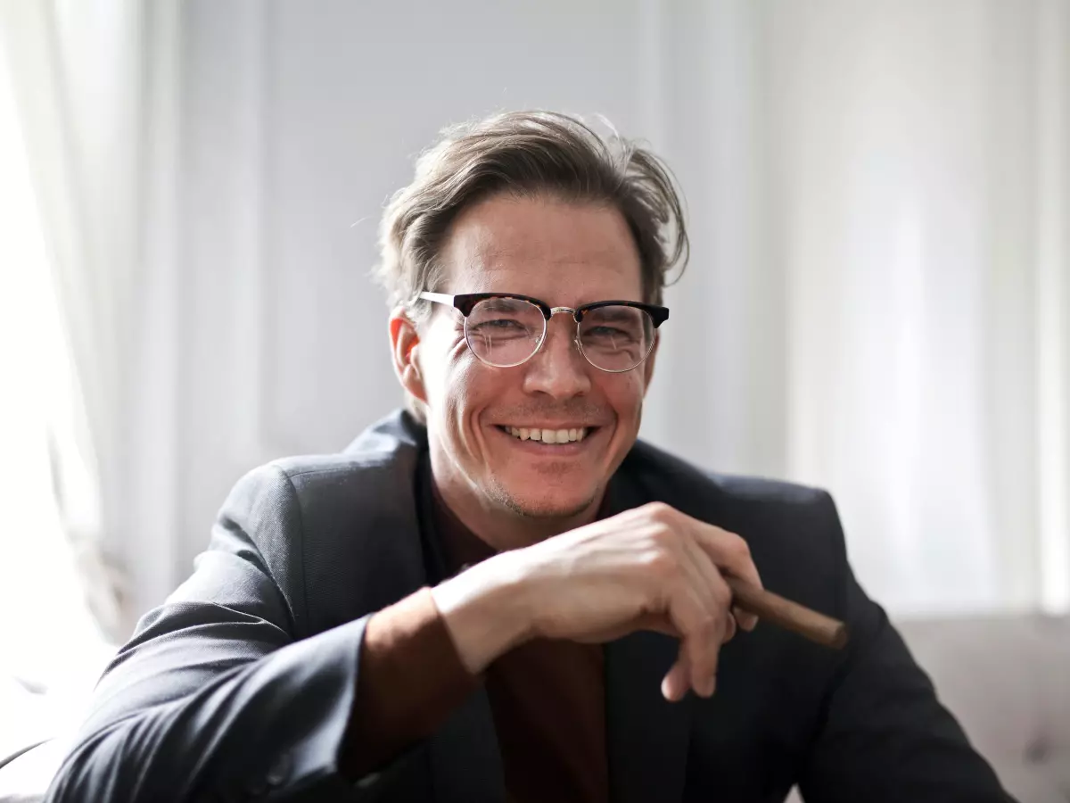 A man in a suit and glasses is sitting on a couch, smiling.